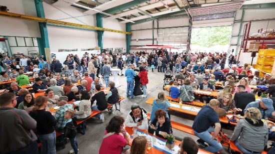 In einem Lagerhalle sind viele Festgarnituren aufgestellt. Menschen sitzen zusammen und unterhalten sich.
