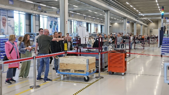 In einer Industriehalle stehen Menschen vor einem Absperrband. Ein Mann zeigt auf Maschinenteile.