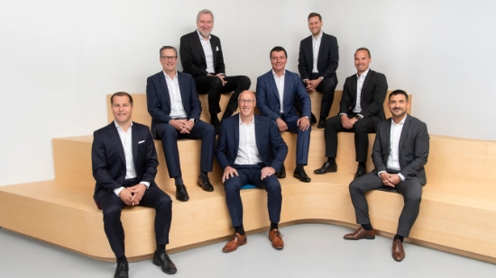 Group photo of the management: Eight men in suits sit on a wooden seating element. They are arranged in three rows.