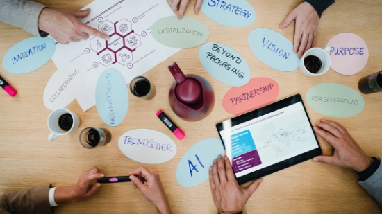 Several hands point to a table with labeled cards, diagrams, pens, coffee cups, and a tablet.