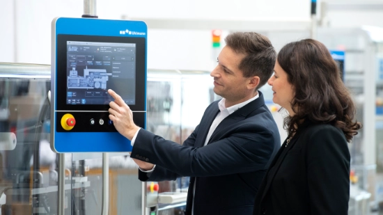 Two people in business attire discussing machine functions on a Uhlmann touch screen in an industrial environment.