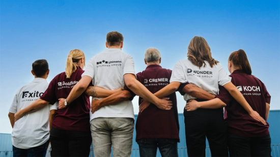 Six people standing arm in arm, wearing shirts with the company logos of axito, Goldfuss, Uhlmann, CREMER, WONDER, and KOCH.