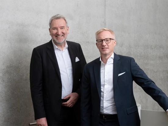 CFO Alexander Schöllhorn (left) and CEO Prof. Dr.-Ing. Matthias Niemeyer standing together indoors in front of a concrete wall