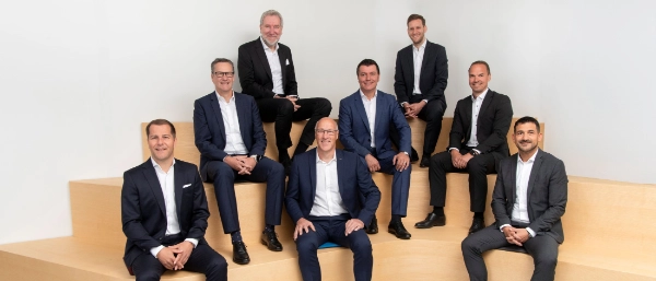 Gruppenfoto der Geschäftsleitung: Acht Männer in Anzügen sitzen auf einem Sitzelement aus Holz. Sie sind in drei Reihen angeordnet.
