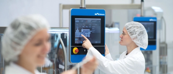Uhlmann employees wearing protective clothing operating an Uhlmann packaging line via a touchscreen and checking blister packaging