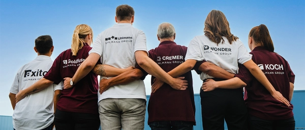 Six people standing arm in arm, wearing shirts with the company logos of axito, Goldfuss, Uhlmann, CREMER, WONDER, and KOCH.