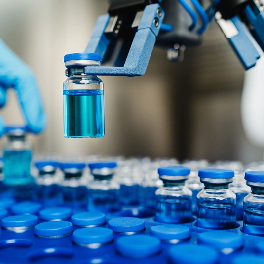Robotic arm holding a small glass vial filled with blue liquid, surrounded by sealed vials arranged in rows.
