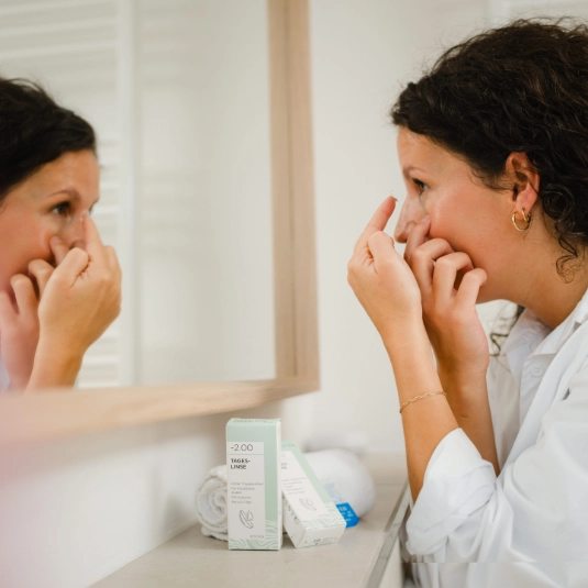 A person applying a contact lense in front of a bathroom mirror, with the product packaging visible.